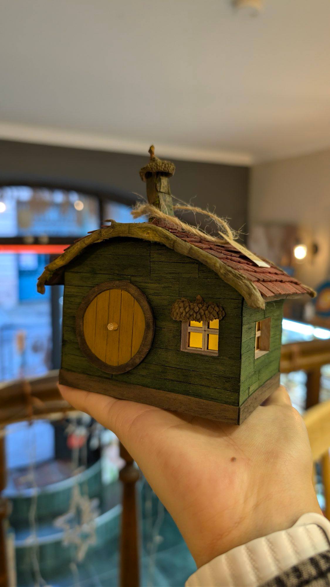 A moss-green cottage with a round yellow door and an acorn cap chimney, held in one hand indoors