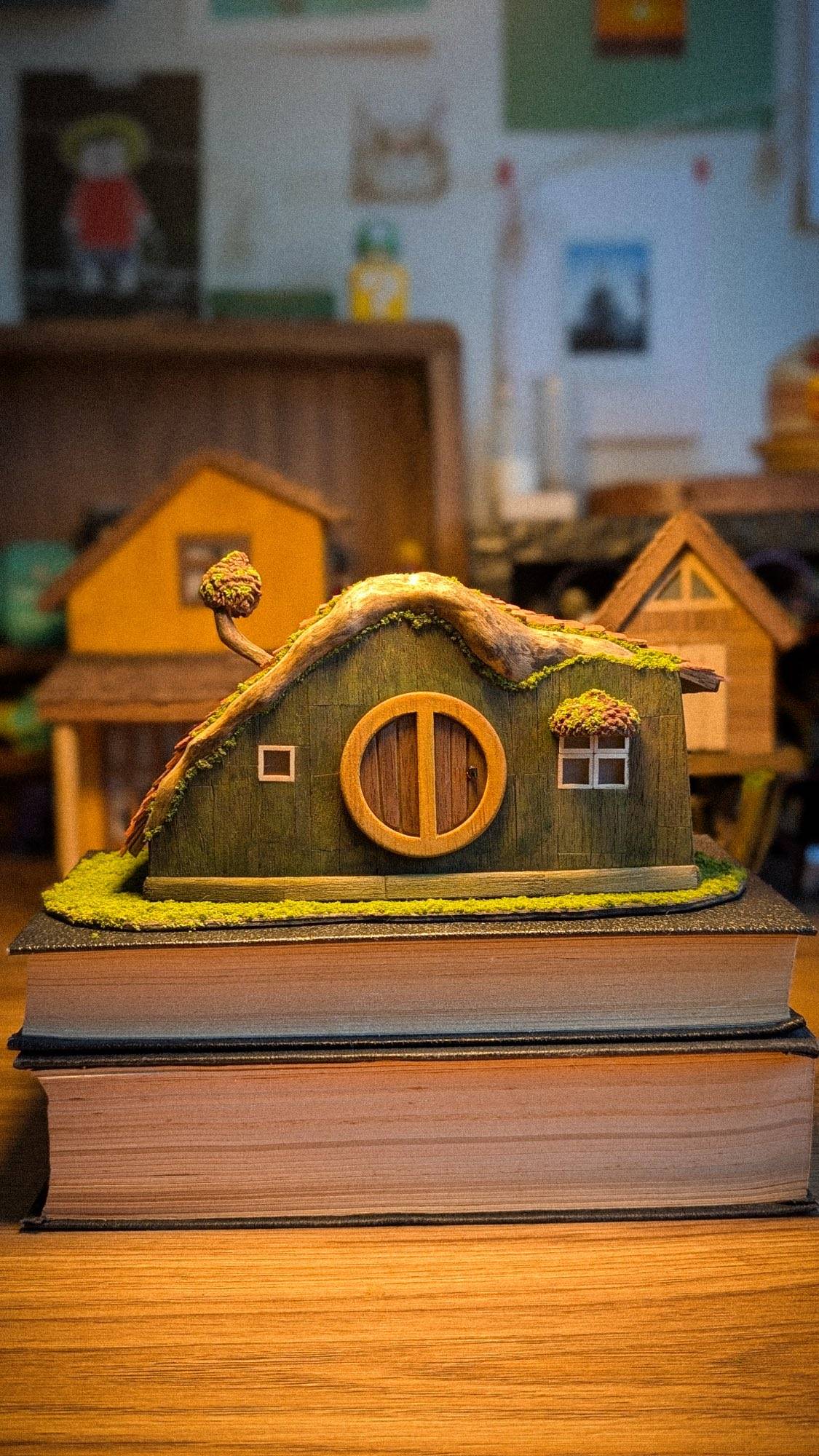 A long green cottage with a round wooden door and a mossy clay roof, sitting on a stack of two hardback books