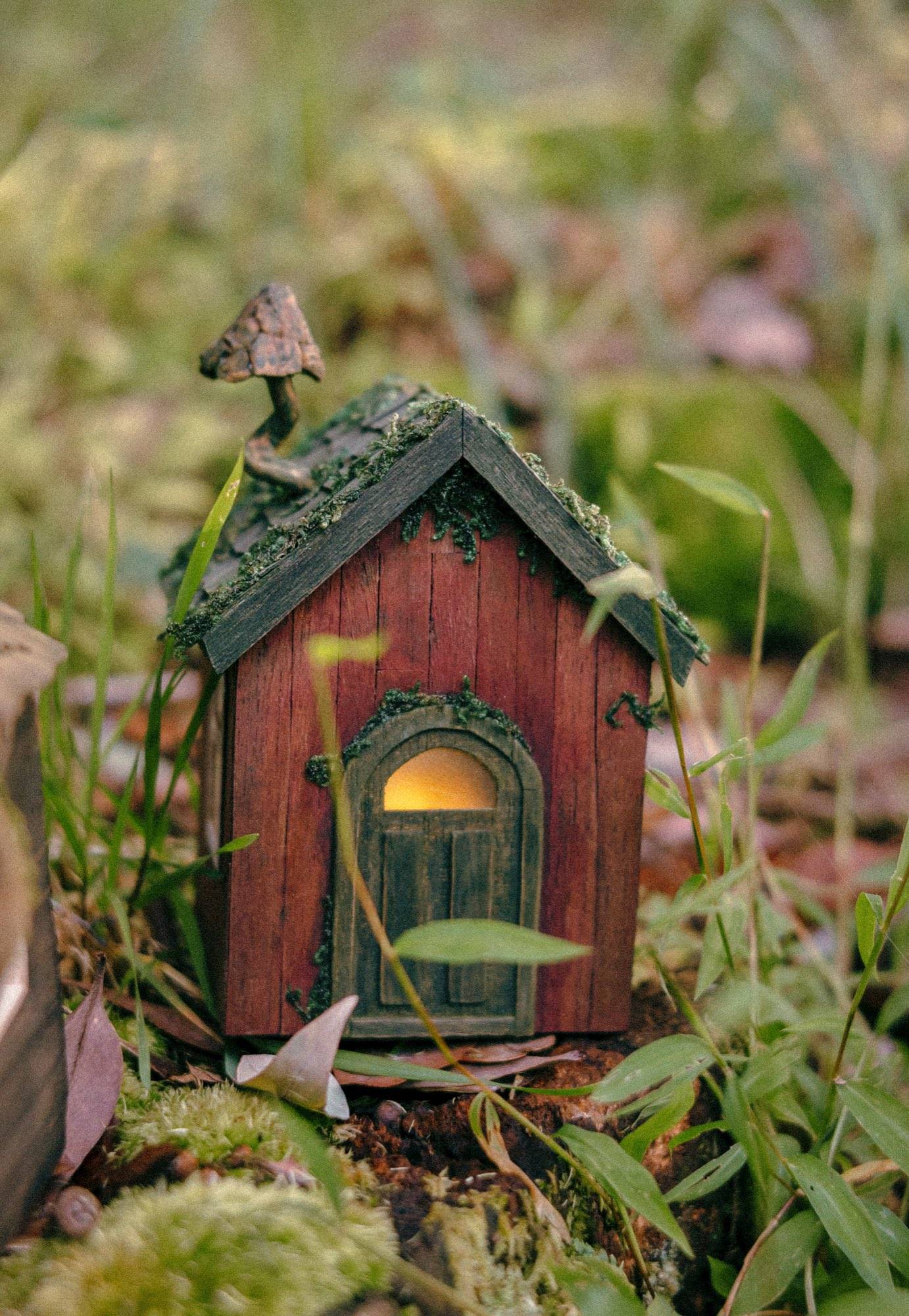 A small red-brown cottage with a moss-covered shingle roof and a deep arched door lit warm from inside, standing in grass