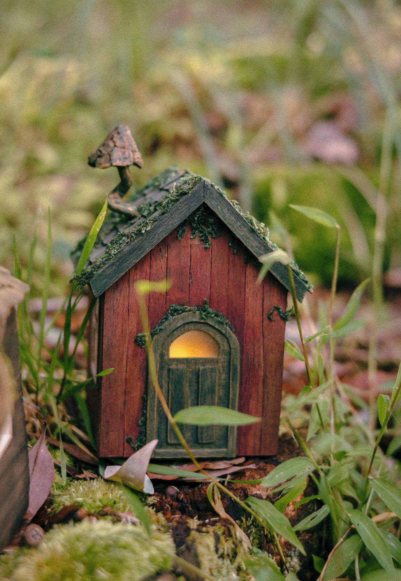 A small red-brown cottage with a moss-covered shingle roof and a deep arched door lit warm from inside, standing in grass