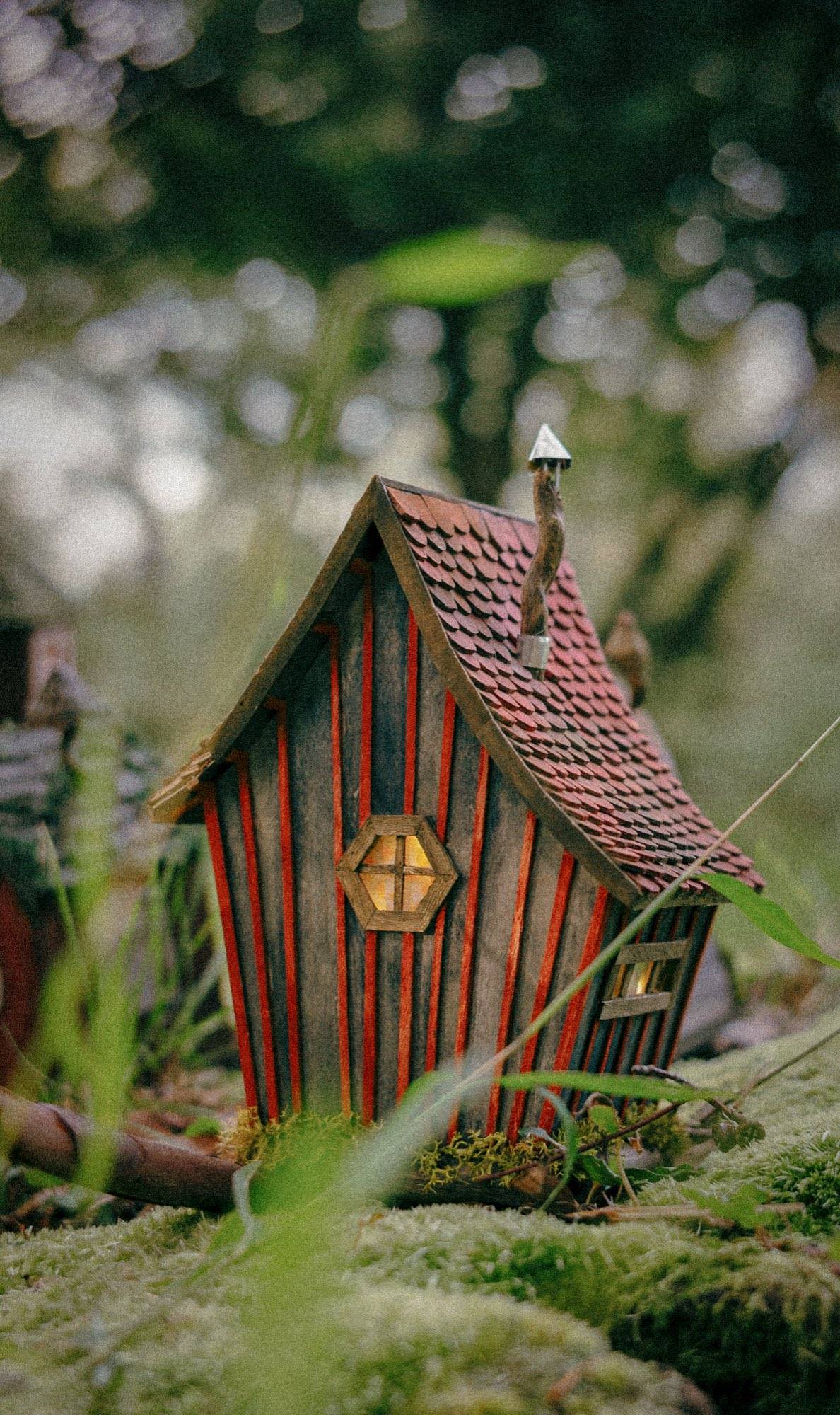 A narrow striped cottage with a scalloped clay-red tile roof and a hexagonal window lit warm from inside, standing on moss