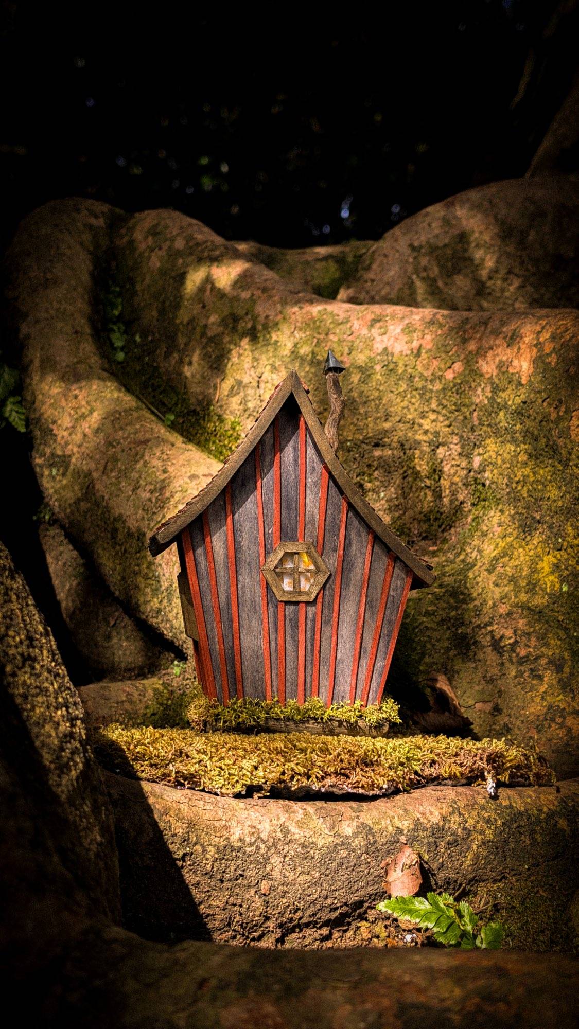 The same cottage between tree roots after dark, the hexagonal window the only light in the frame