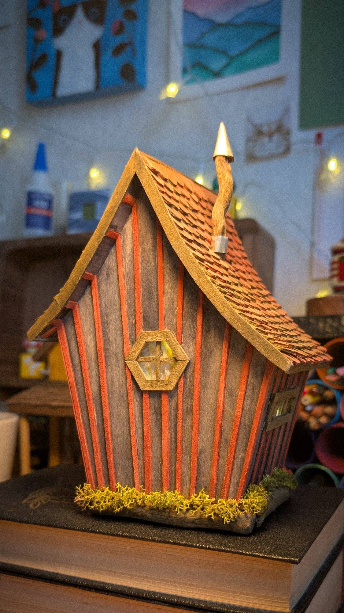 A finished striped cottage standing on a book at the workbench, its window lit, with glue bottles, drawings pinned to the wall and a string of fairy lights behind it